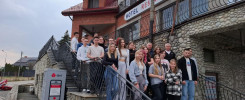 Grupa osób stoi na schodach przed budynkiem z napisem "HOTEL RESTAURACJA". Obok znajduje się paczkomat DPD.