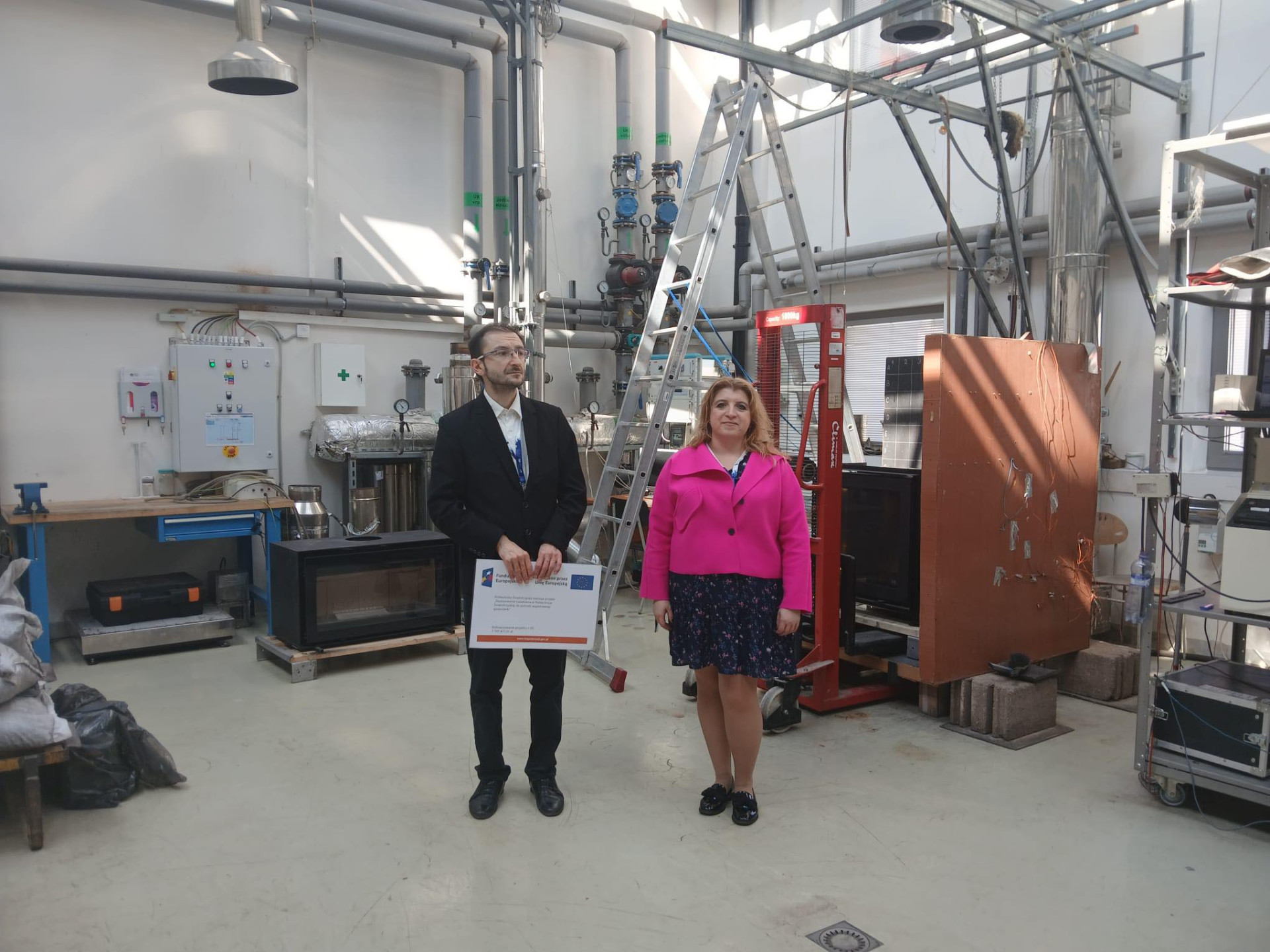 Mężczyzna i kobieta stoją w laboratorium. W tle widoczne są rury, drabina i sprzęt badawczy.