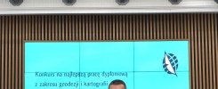 Dwóch mężczyzn stoi przed ekranem z napisem "Konkurs na najlepszą pracę dyplomową z zakresu geodezji i kartografii".