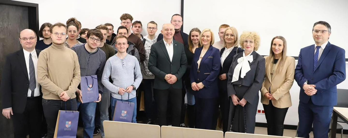 Grupa osób, w tym mężczyźni i kobiety, stoi razem. W tle widać białą ścianę i drzwi.
