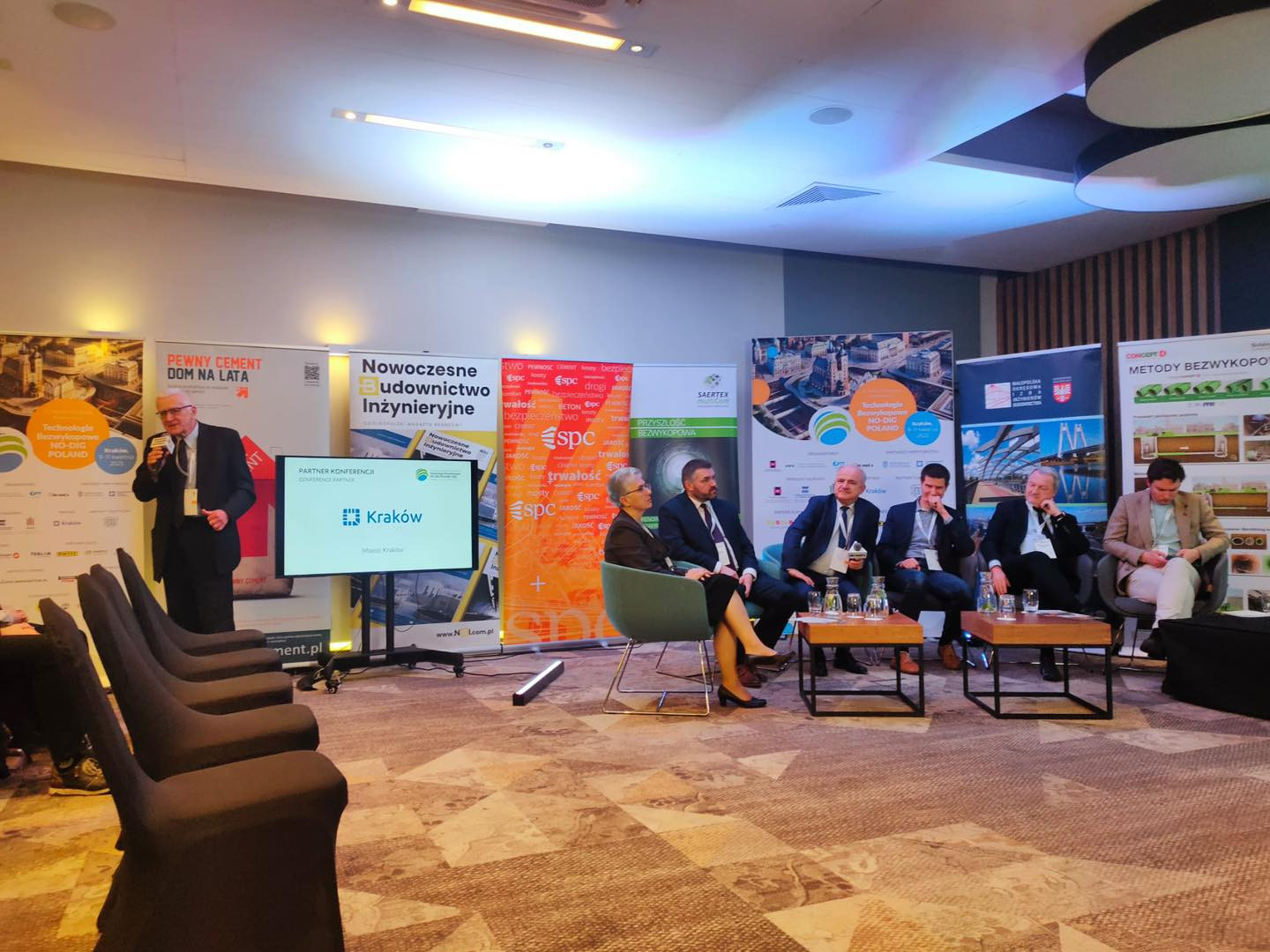 Panel dyskusyjny na konferencji budowlanej. Uczestnicy siedzą przy stołach, jeden mężczyzna przemawia.