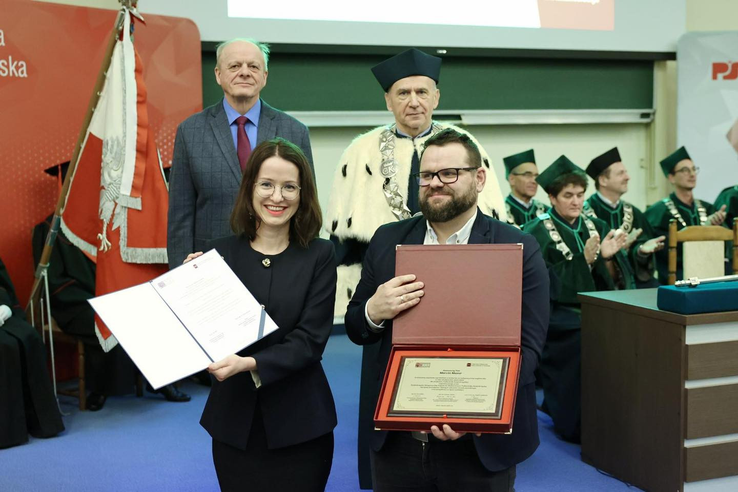 Kobieta i mężczyzna odbierają dyplomy i nagrodę. W tle stoją osoby w togach akademickich.