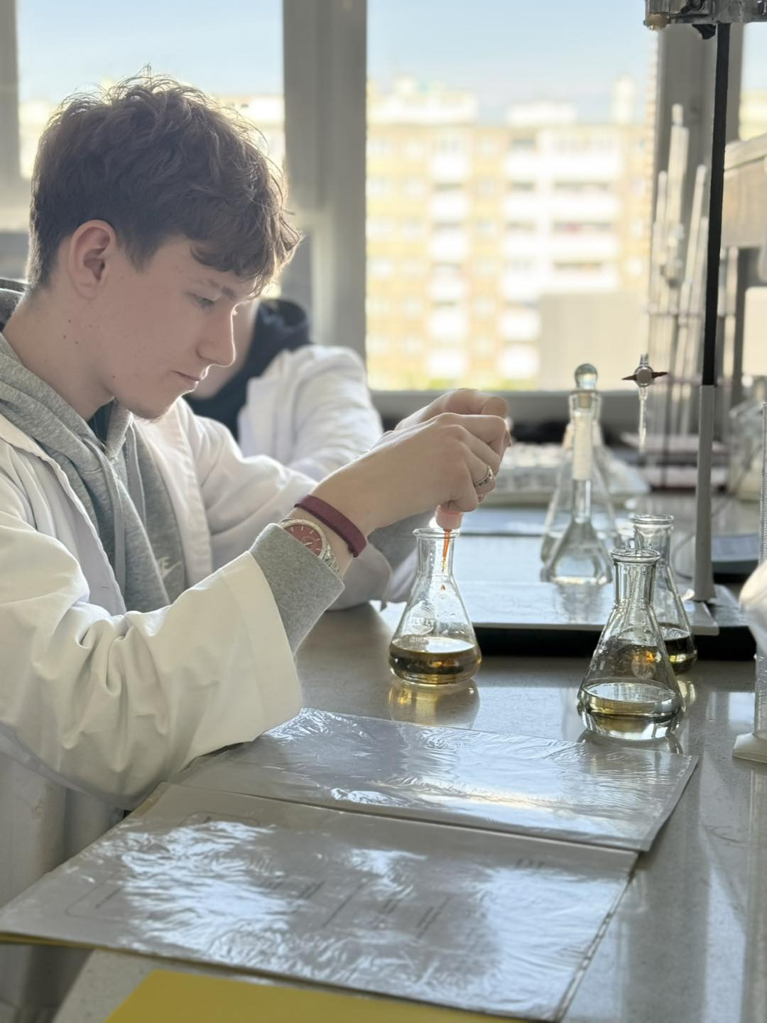 Młody mężczyzna w fartuchu laboratoryjnym wykonuje eksperyment. Kluczowe obiekty: kolby, płyny, fartuchy.