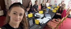 Grupa młodych ludzi siedzi przy stole w sali konferencyjnej. Na stole projektor, laptopy i żółte kartki.