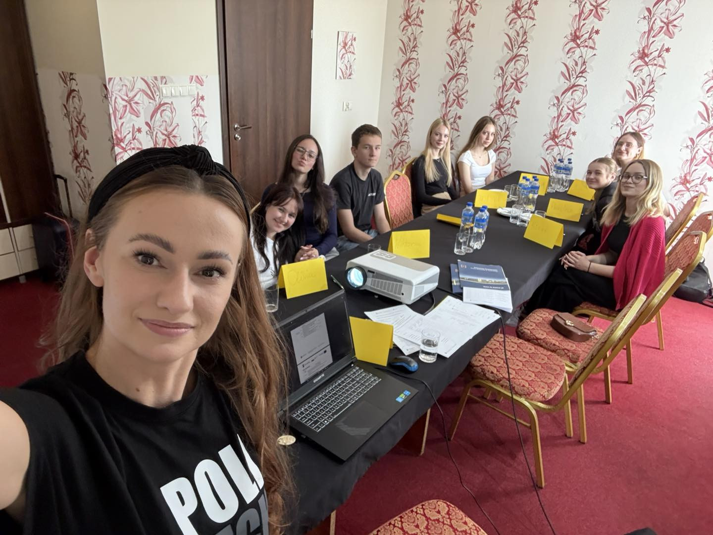 Grupa młodych ludzi siedzi przy stole w sali konferencyjnej. Na stole projektor, laptopy i żółte kartki.