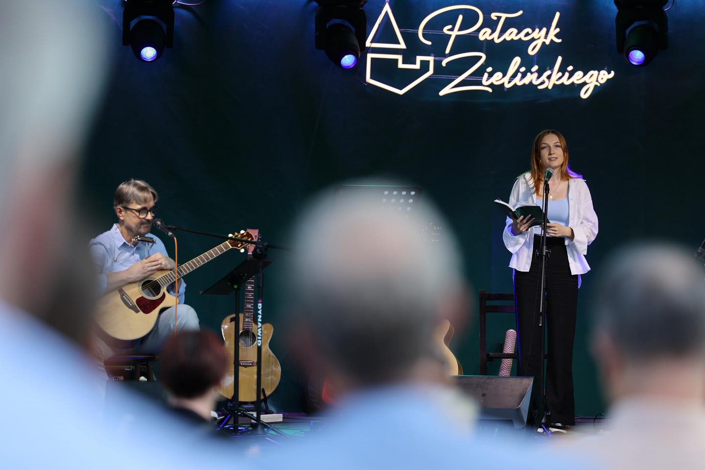 Mężczyzna gra na gitarze, kobieta czyta z książki. W tle neonowy napis "Pataczyk Zielińskiego". Koncert.