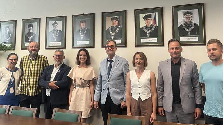 Grupa osób pozuje do zdjęcia na tle portretów. W kadrze: 4 kobiety, 4 mężczyzn, portrety.