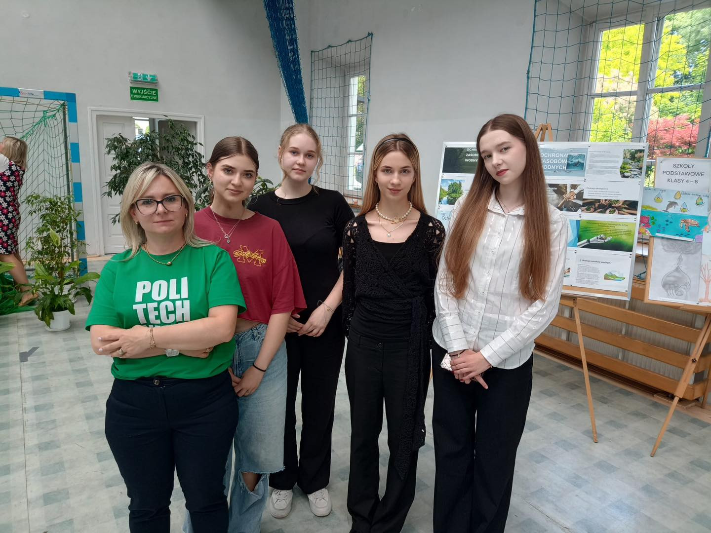 Grupa osób stoi przed tablicami edukacyjnymi. Kluczowe obiekty: kobiety, tablice, rośliny.