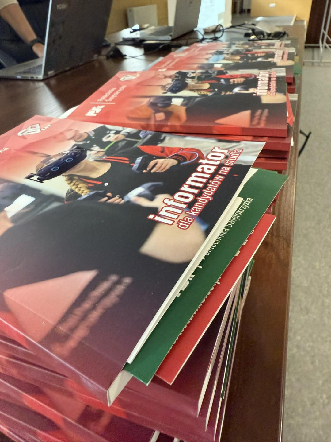 Stos informatorów "Informator dla kandydatów na studia". Kluczowe obiekty: informatory, stół, laptopy.