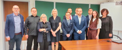 Grupa osób stoi przed zieloną tablicą z polskimi godłami. W kadrze widać biurko.
