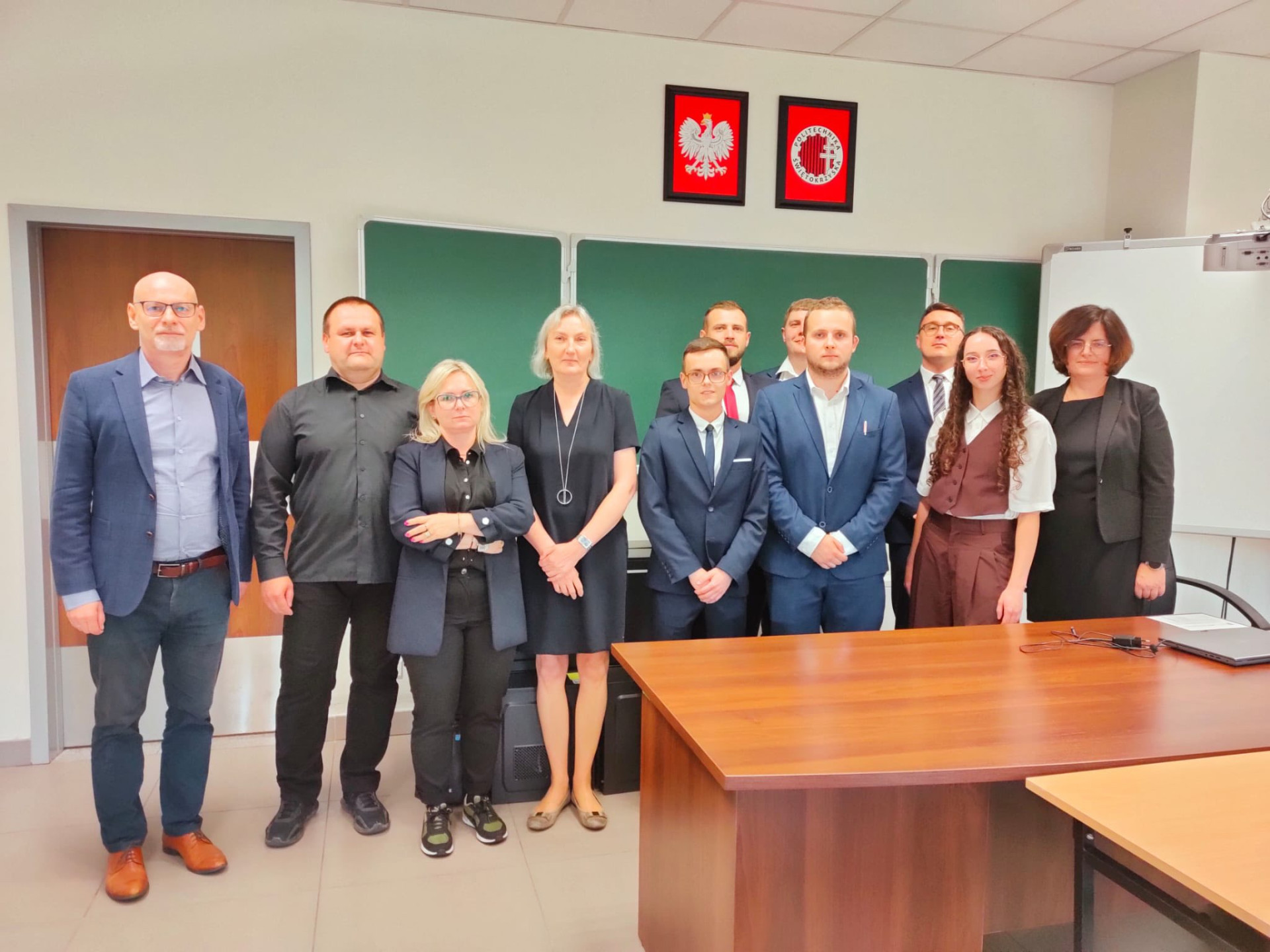 Grupa osób stoi przed zieloną tablicą z polskimi godłami. W kadrze widać biurko.