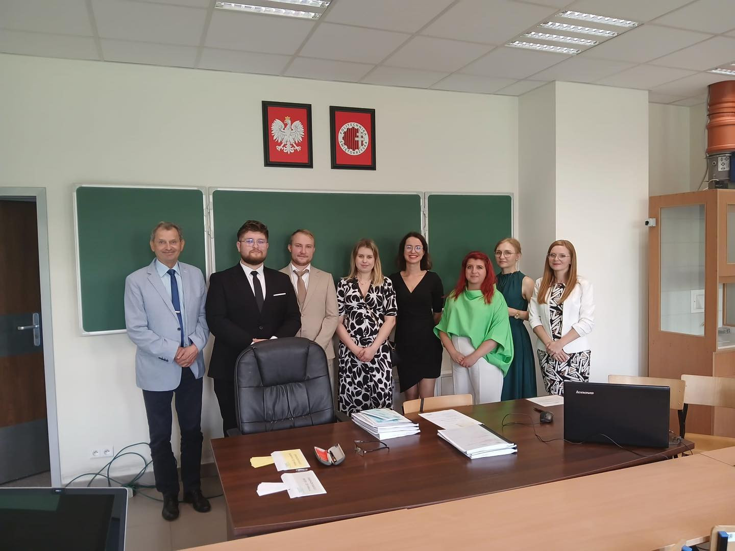 Osiem osób stoi przed zieloną tablicą. Na ścianie widnieją godła. Kontekst: sala wykładowa.