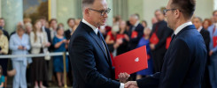 Dwóch mężczyzn w garniturach ściska sobie dłonie. Jeden z nich trzyma czerwony folder. W tle stoi grupa ludzi.