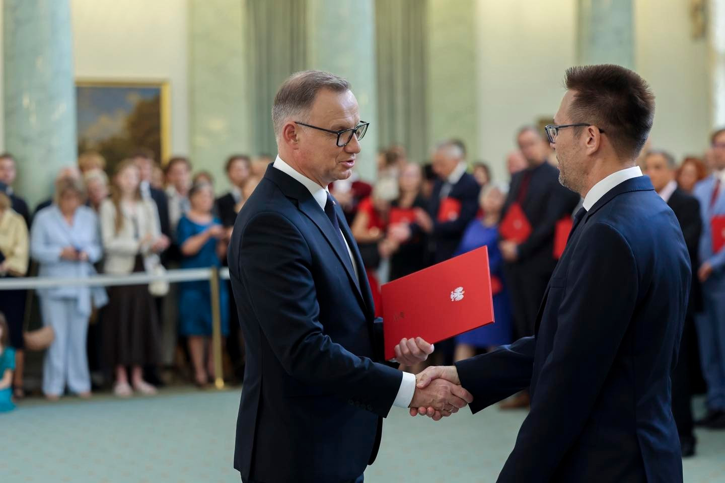 Dwóch mężczyzn w garniturach ściska sobie dłonie. Jeden z nich trzyma czerwony folder. W tle stoi grupa ludzi.