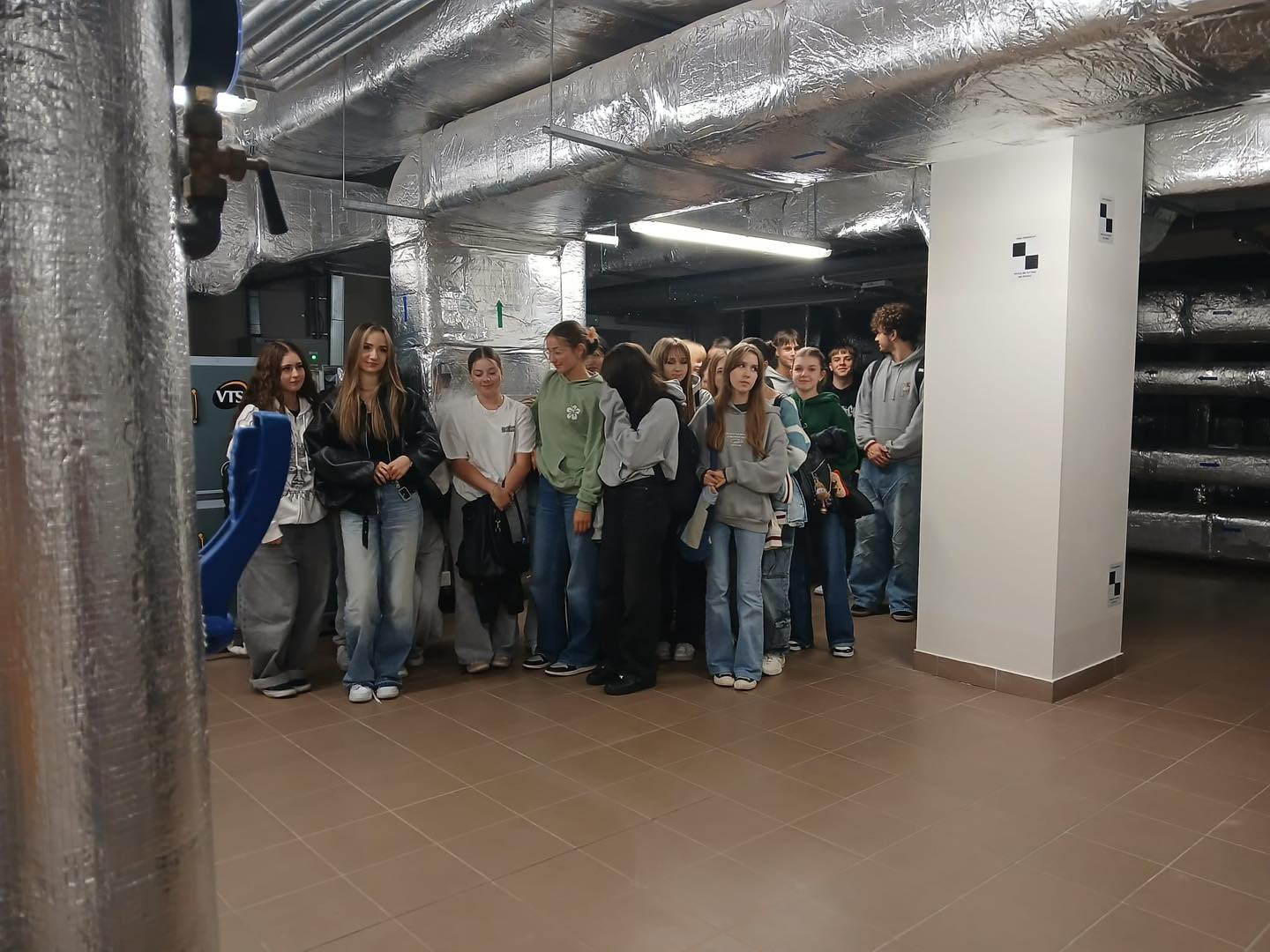 Grupa młodzieży stoi w pomieszczeniu technicznym z rurami i izolacją. Widać kolumnę i urządzenie z napisem VTS.