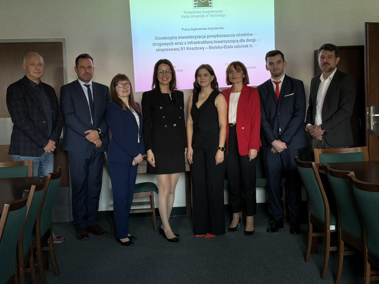 Grupa osób stoi przed tablicą z tekstem. Widać mężczyzn i kobiety w strojach formalnych.