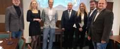 Grupa siedmiu osób stoi przed ekranem projekcyjnym. W sali znajdują się stoły i krzesła.