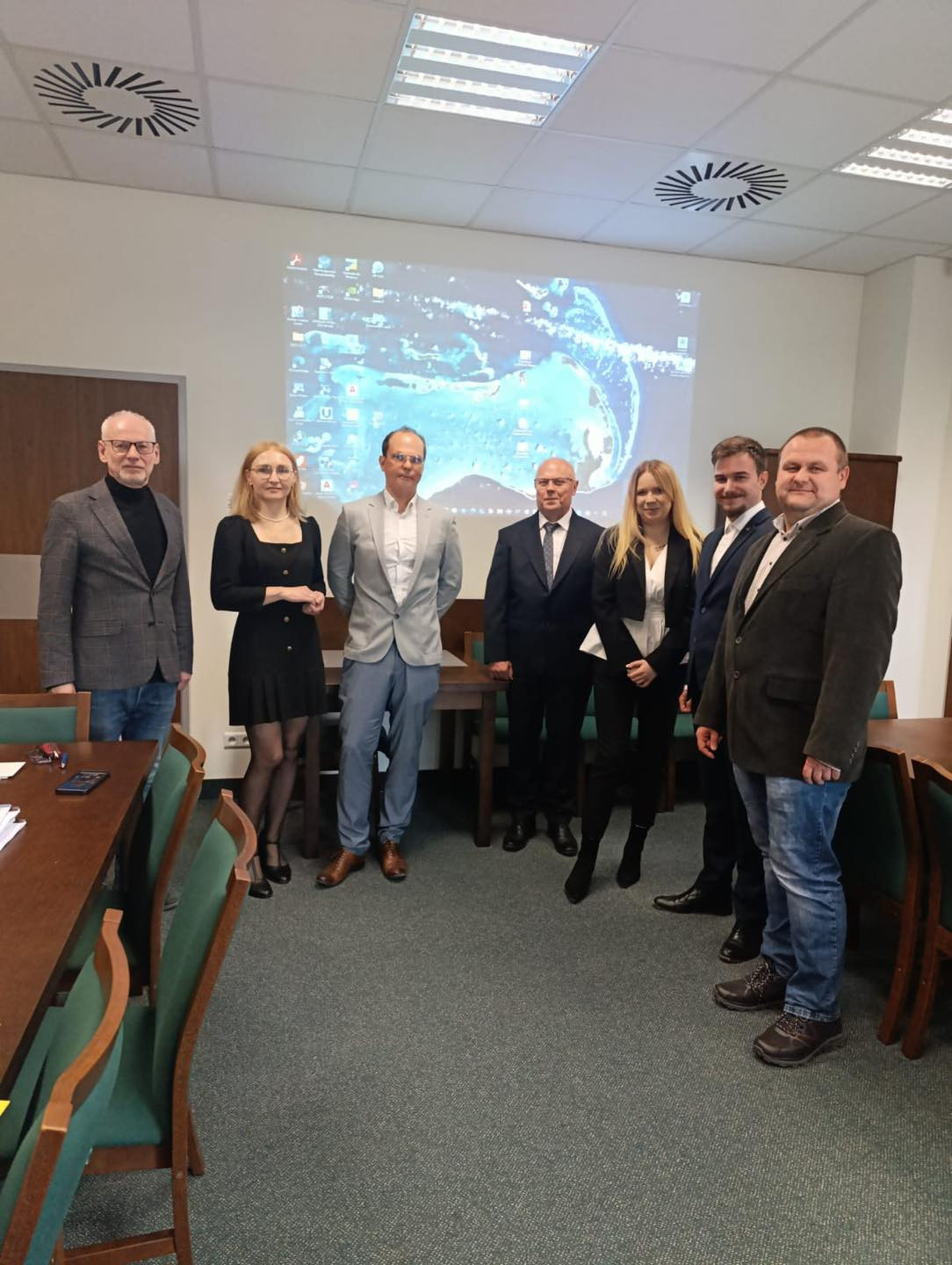 Grupa siedmiu osób stoi przed ekranem projekcyjnym. W sali znajdują się stoły i krzesła.