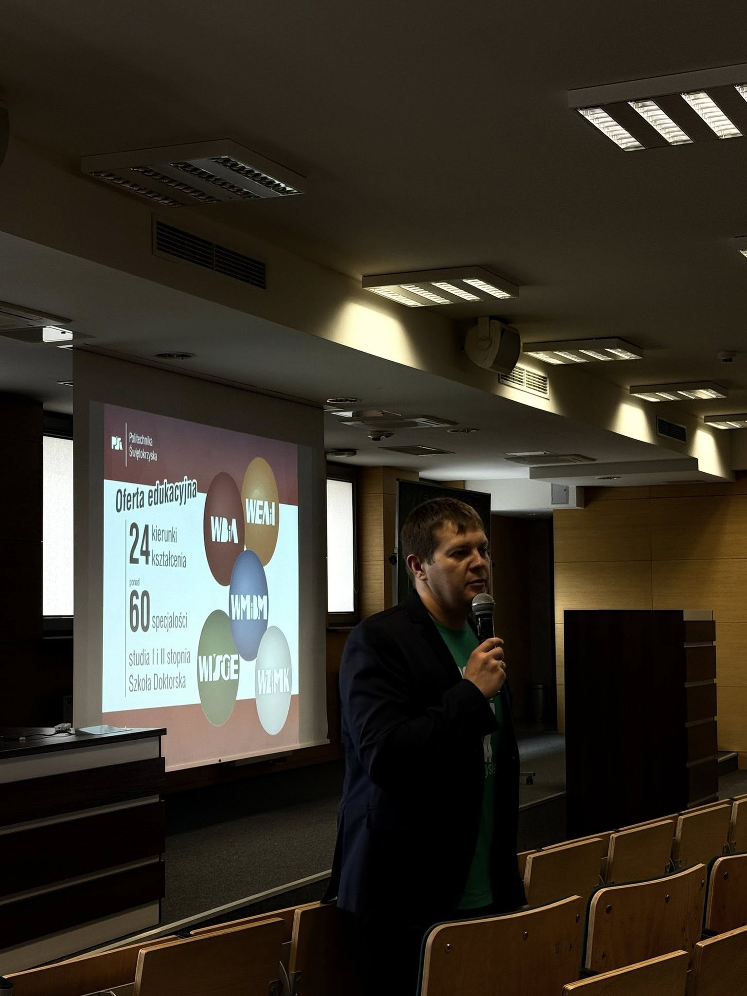 Mężczyzna z mikrofonem przemawia na tle prezentacji o ofercie edukacyjnej Politechniki Świętokrzyskiej.