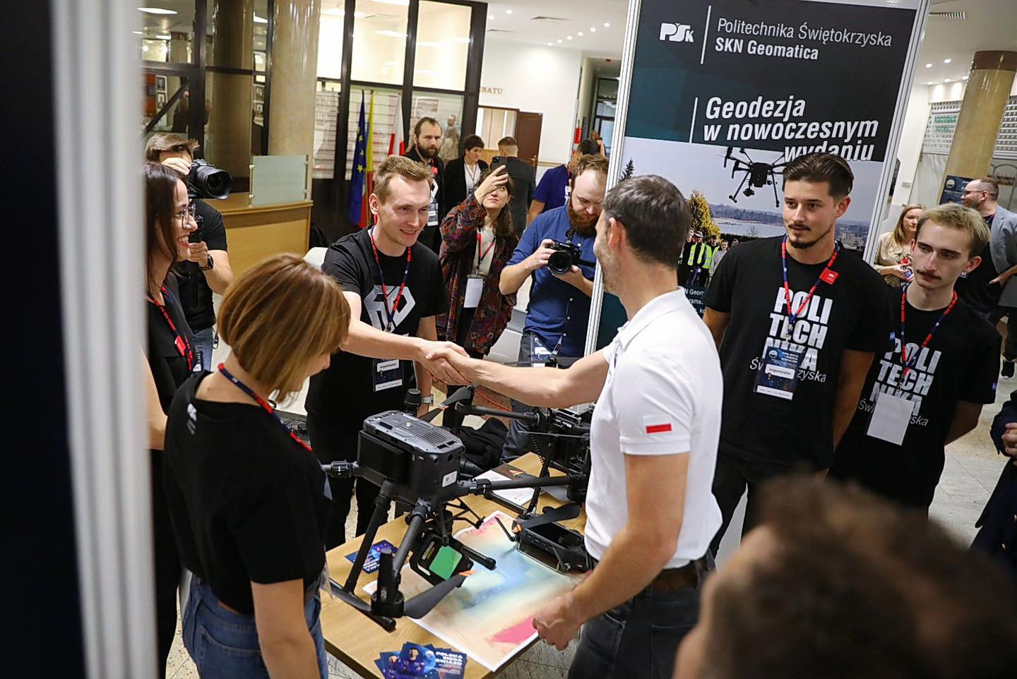 Ludzie przy stoisku z dronem, jeden mężczyzna ściska dłoń drugiemu. Kontekst: wydarzenie naukowe lub targi.