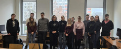 Grupa osób stoi w sali lekcyjnej. Widać okna, biurka i krzesła.
