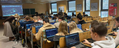 Sala wykładowa z uczniami pracującymi na laptopach. Na ekranie projekcyjnym widoczny jest schemat.