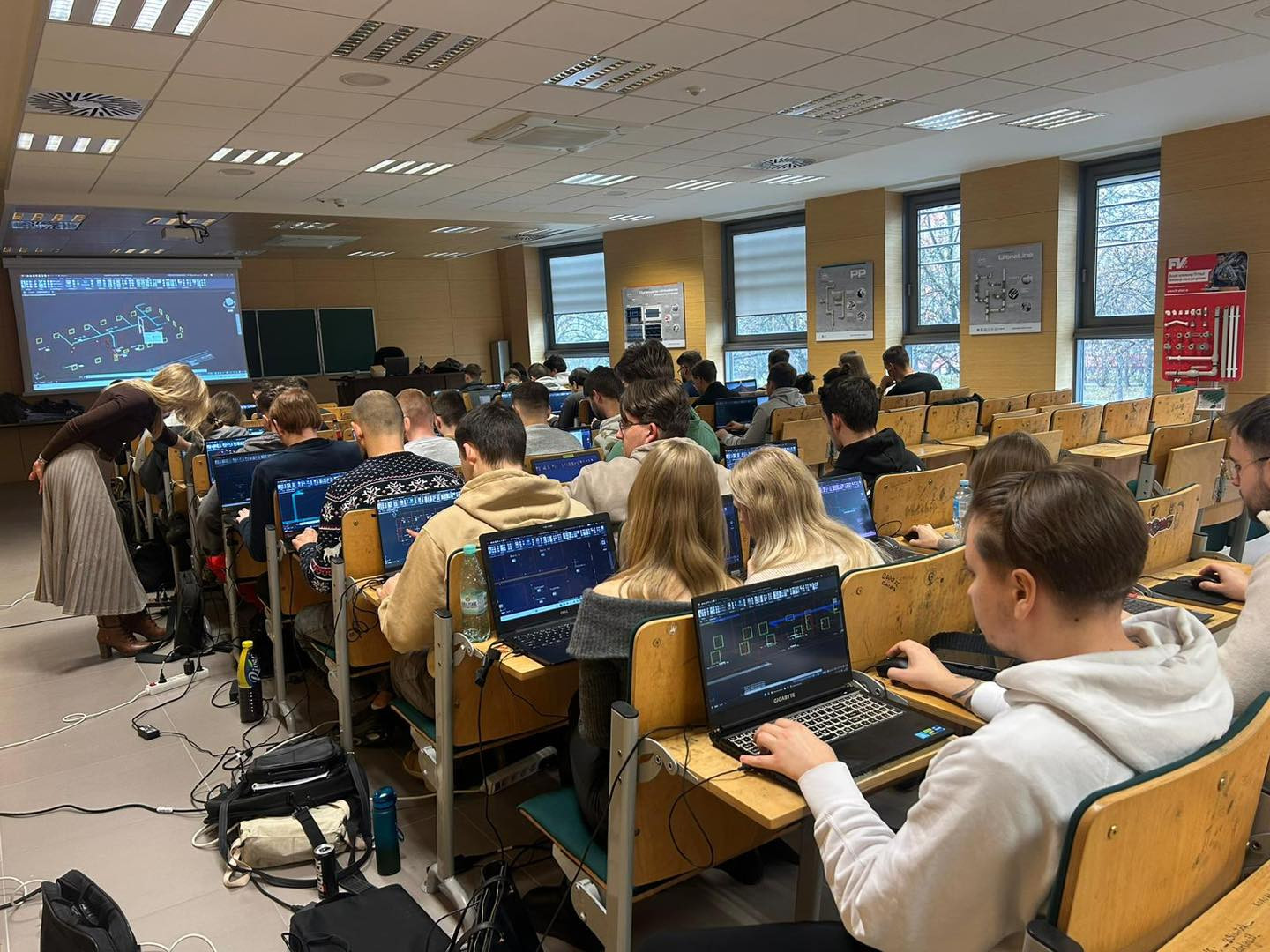 Sala wykładowa z uczniami pracującymi na laptopach. Na ekranie projekcyjnym widoczny jest schemat.