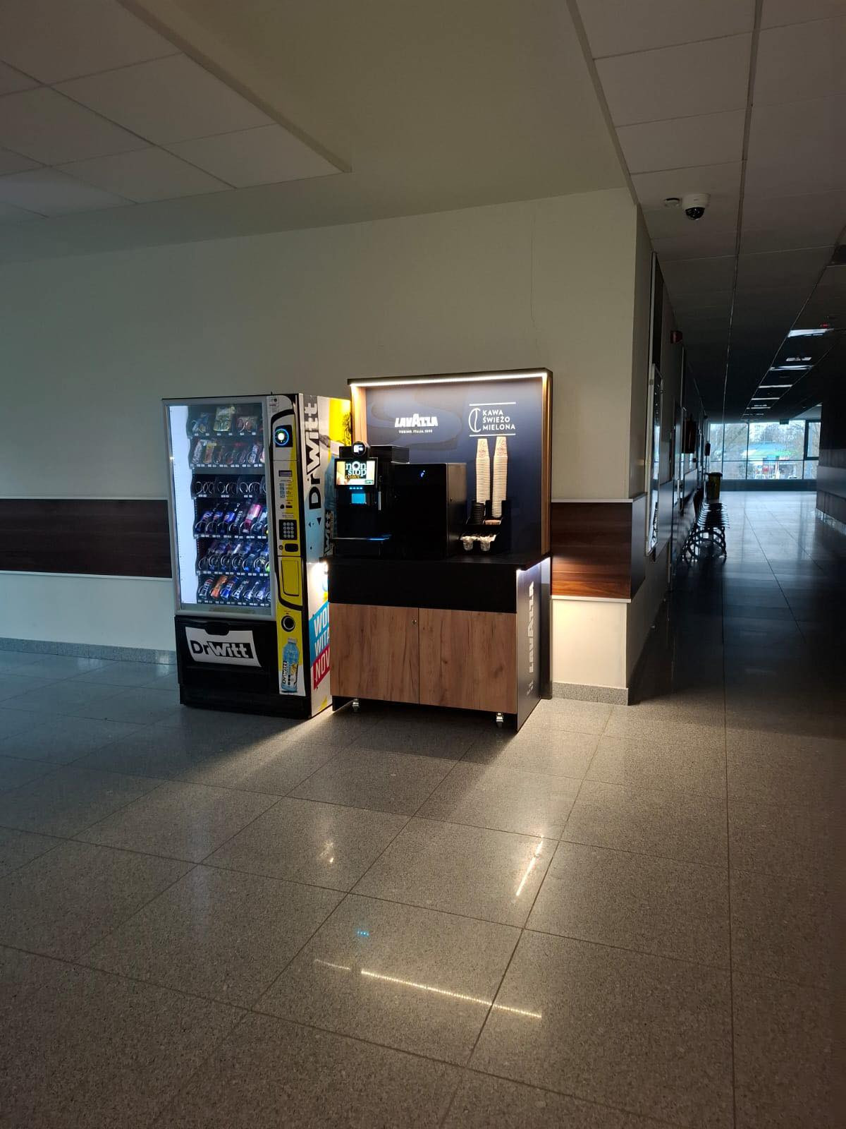 Automat z napojami i ekspres do kawy Lavazza stoją w korytarzu. Kluczowe obiekty: automat, ekspres, Lavazza, DrWitt.