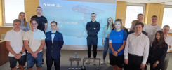 Grupa osób stoi przed ekranem z logo firmy i napisem. Wszyscy są ubrani w stroje casualowe lub formalne.