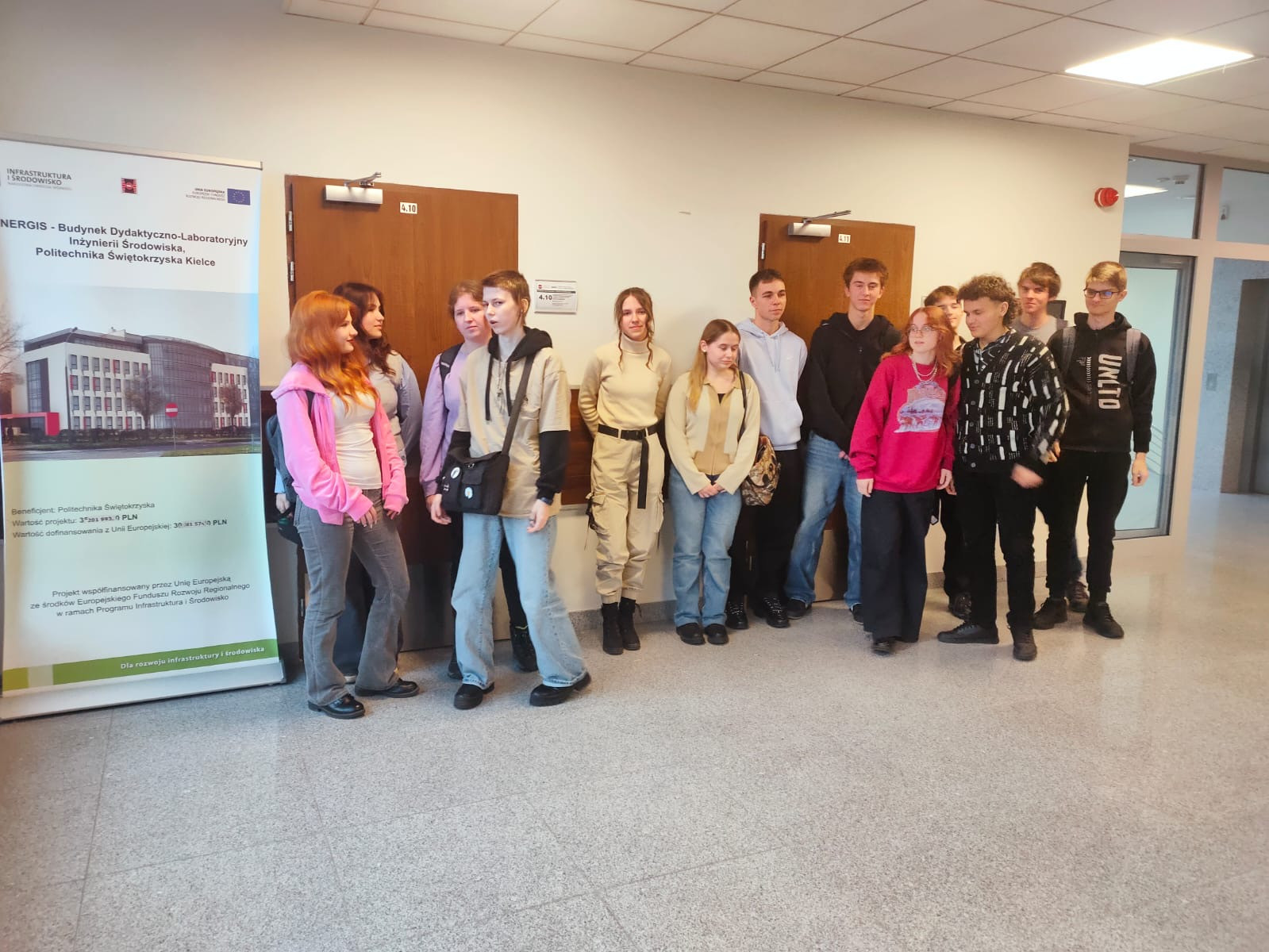 Grupa młodych ludzi stoi przed drzwiami w korytarzu. W tle baner informacyjny.
