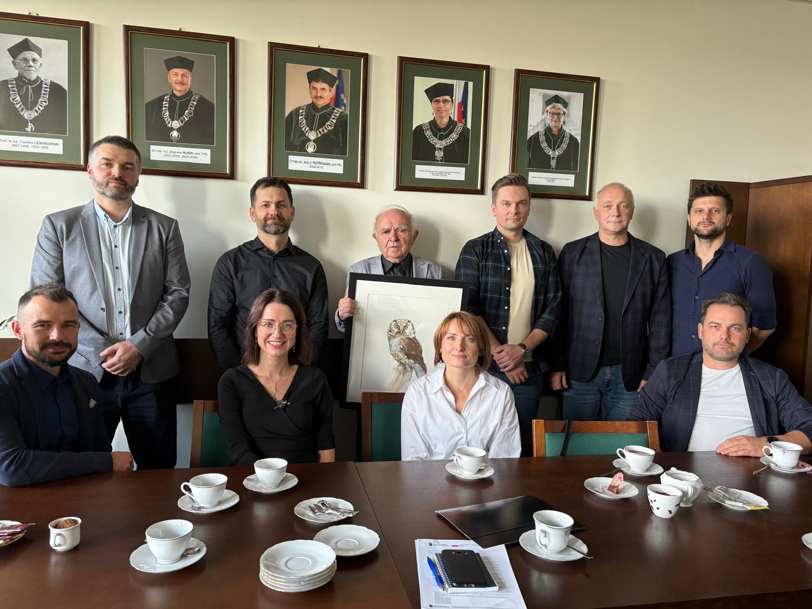 Grupa osób pozuje do zdjęcia w pomieszczeniu z portretami na ścianie. Kluczowe obiekty: ludzie, portrety, stół z filiżankami.