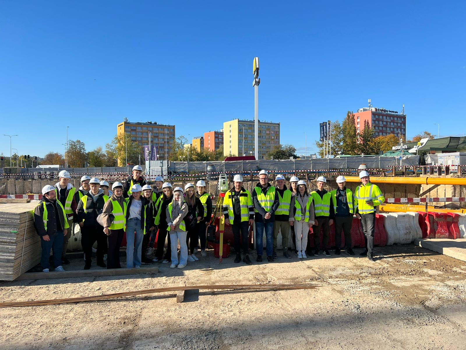 Grupa osób w kaskach i kamizelkach odblaskowych stoi na placu budowy. W tle budynki mieszkalne i McDonald's.