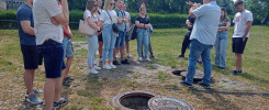Grupa ludzi stoi wokół otwartych włazów. W tle budynek i drzewa.