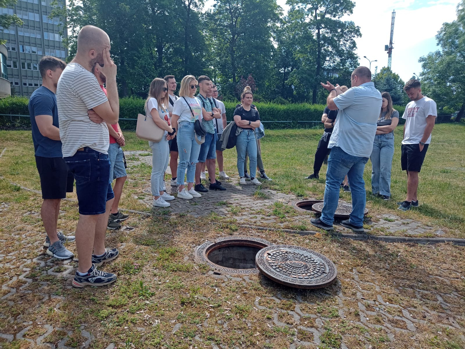 Grupa ludzi stoi wokół otwartych włazów. W tle budynek i drzewa.