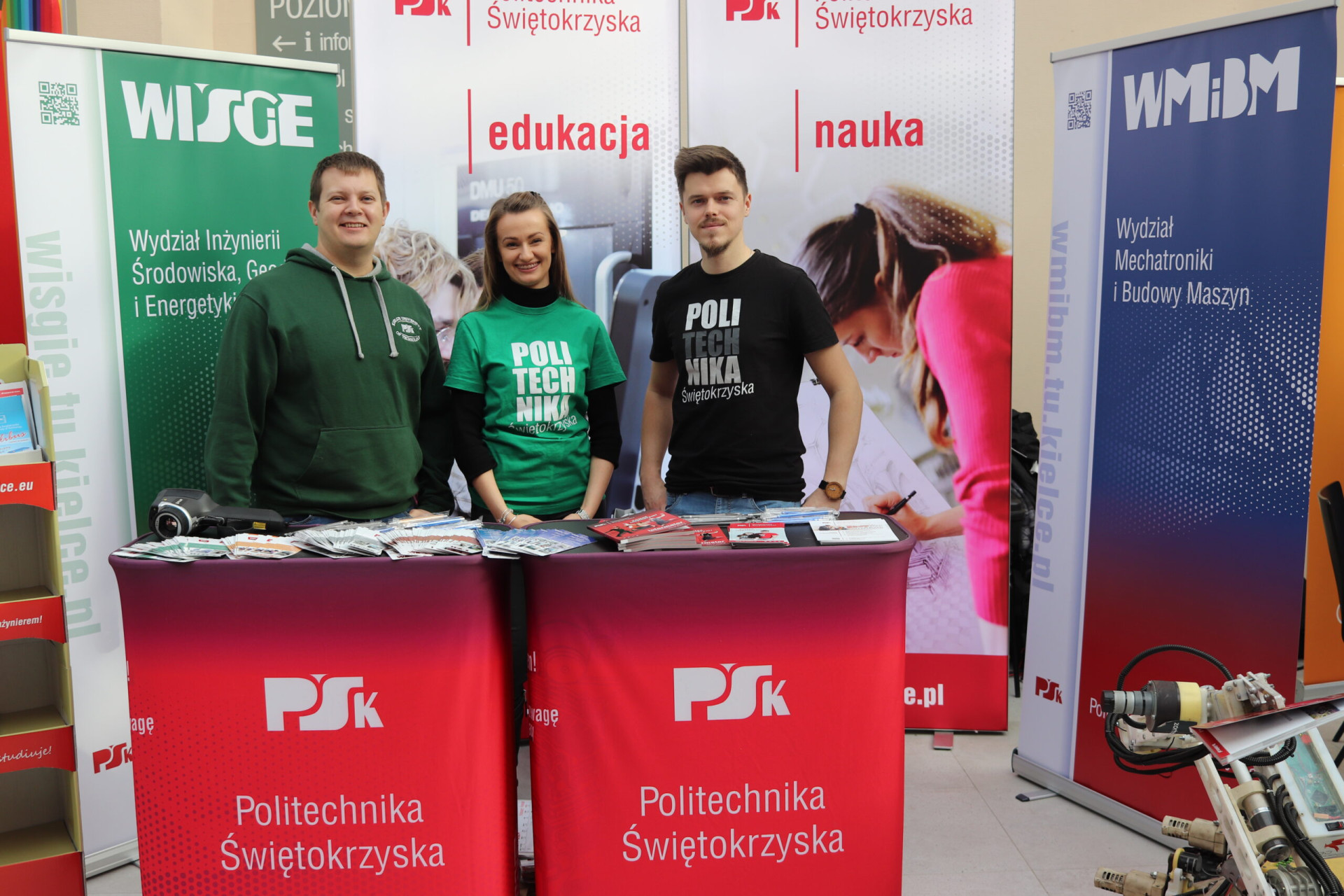 Trzy osoby stoją przy stoiskach promocyjnych Politechniki Świętokrzyskiej. W tle banery uczelni i robot.