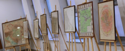 Wystawa map historycznych, prezentująca plany miast i regionów. Kluczowe obiekty: mapy, sztalugi. Kontekst: wystawa, galeria.