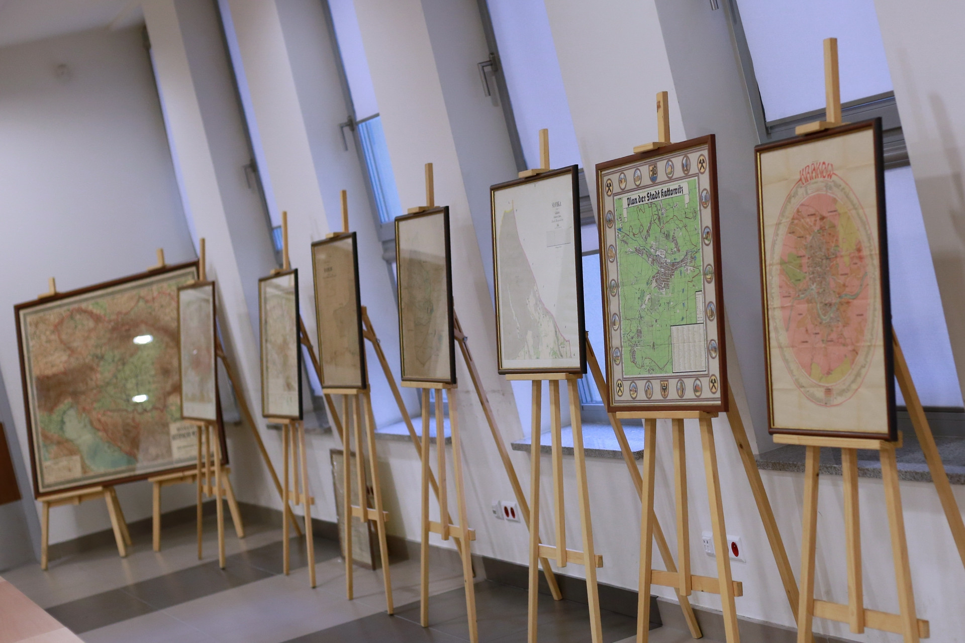 Wystawa map historycznych, prezentująca plany miast i regionów. Kluczowe obiekty: mapy, sztalugi. Kontekst: wystawa, galeria.
