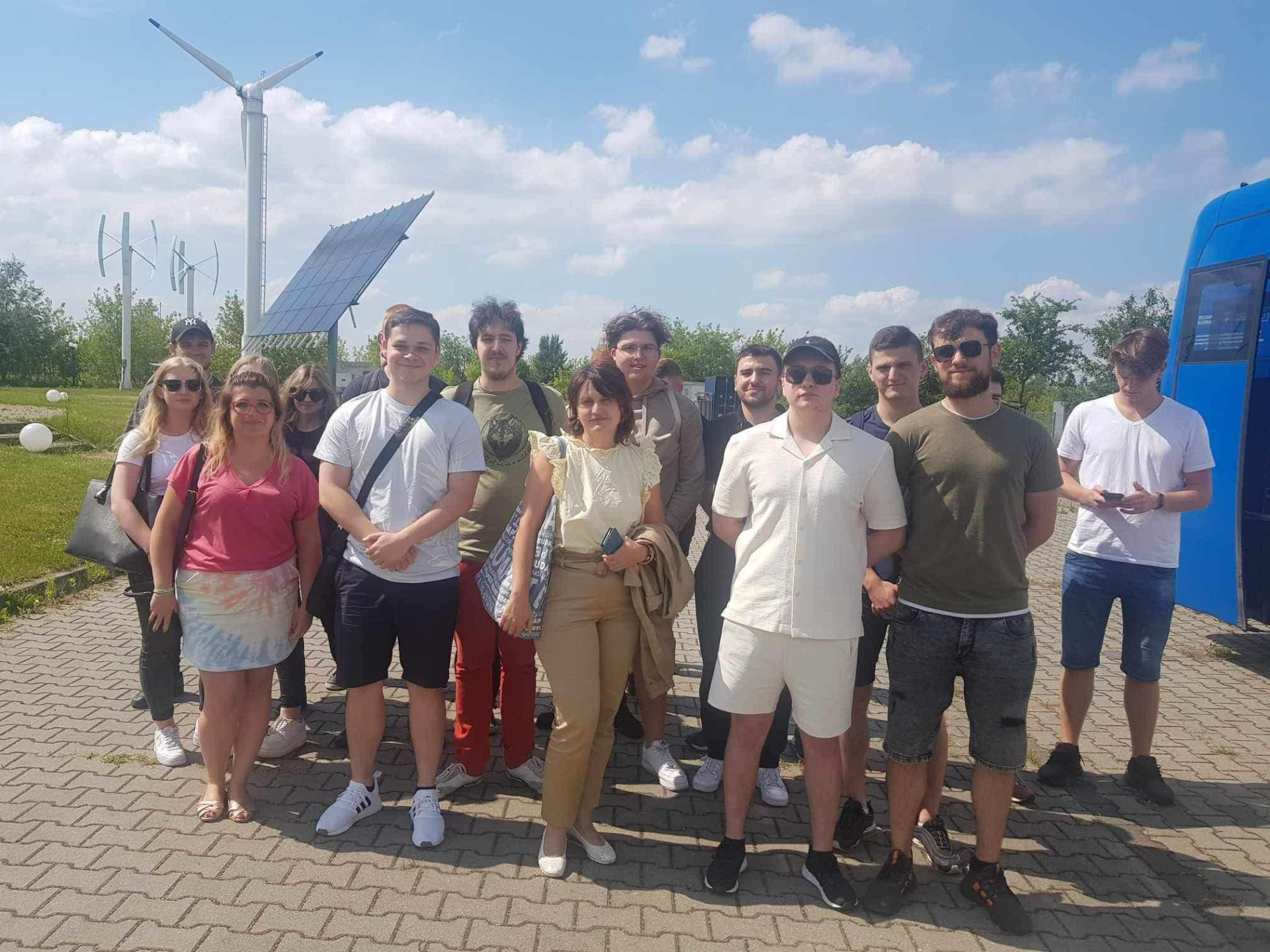 Grupa ludzi stoi przed turbinami wiatrowymi i panelem słonecznym. W tle niebieski pojazd.