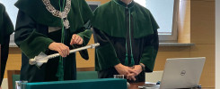 Dwóch mężczyzn w togach akademickich stoi przy stole. Na stole znajduje się laptop, szklanka i ozdobny przedmiot.
