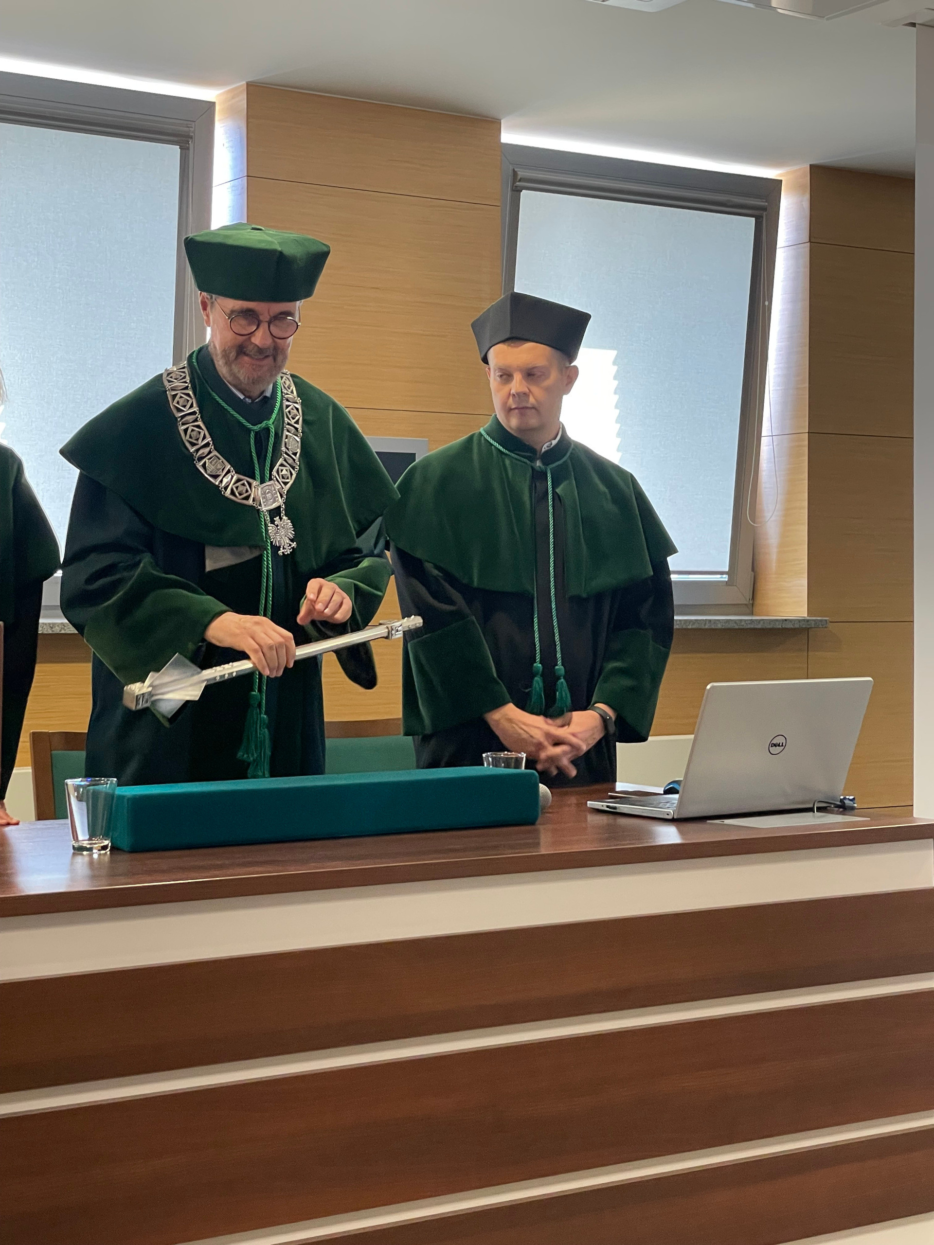 Dwóch mężczyzn w togach akademickich stoi przy stole. Na stole znajduje się laptop, szklanka i ozdobny przedmiot.