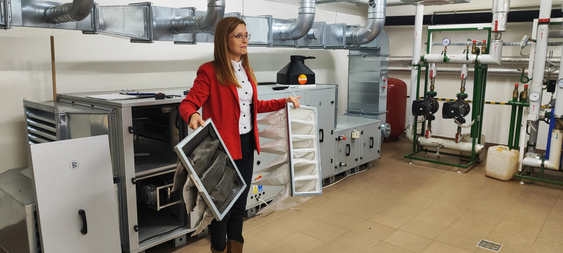 Kobieta w czerwonej marynarce pokazuje brudny i nowy filtr powietrza. W tle widoczne są systemy wentylacyjne i hydrauliczne.