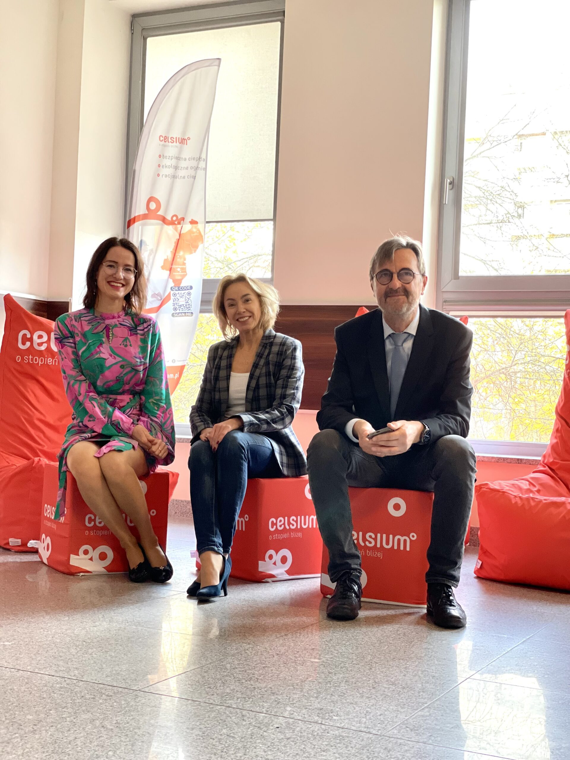 Trzy osoby siedzą na czerwonych pufach i kostkach z logo "celsium". W tle baner.
