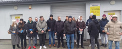 Grupa osób stoi przed budynkiem z tablicami informacyjnymi. Kluczowe obiekty: ludzie, budynek, tablice.