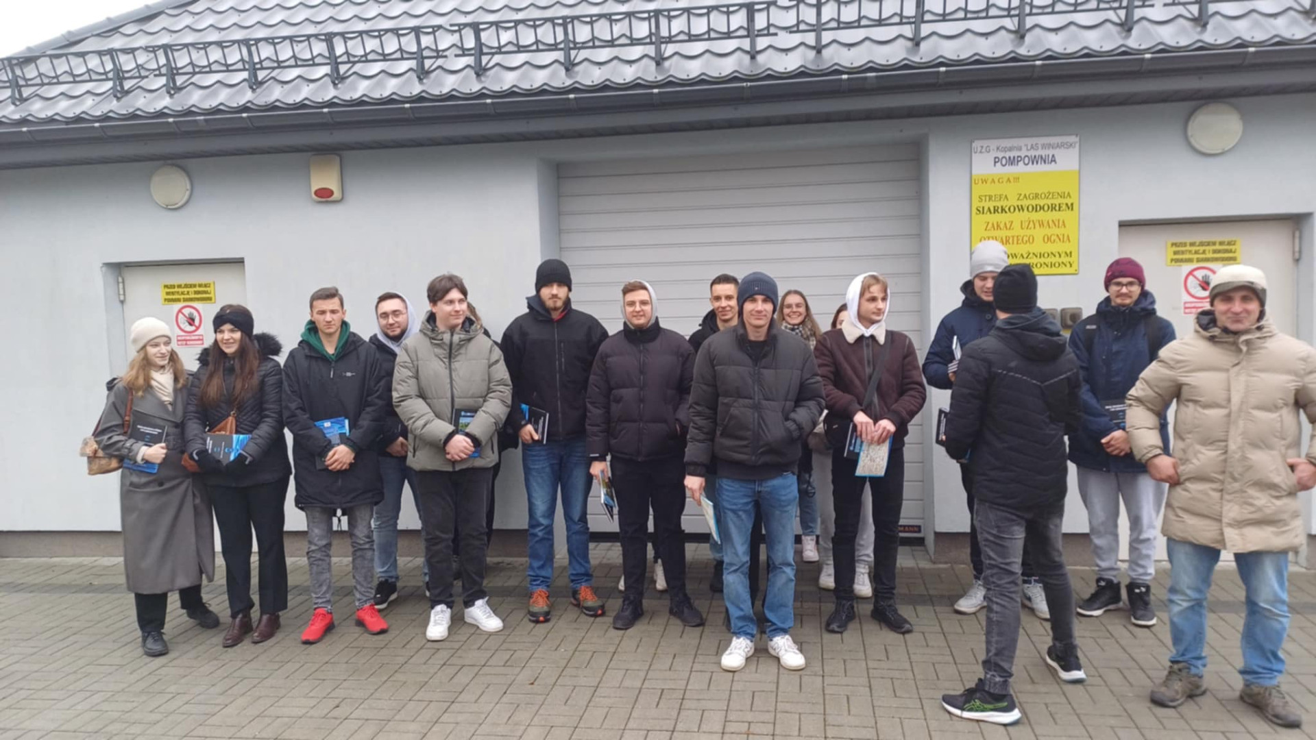 Grupa osób stoi przed budynkiem z tablicami informacyjnymi. Kluczowe obiekty: ludzie, budynek, tablice.
