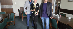 Trzy osoby stoją przy stole w sali konferencyjnej. Kobieta trzyma puchar.