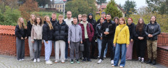 Grupa młodych ludzi stoi na brukowanym placu. W tle budynki i drzewa.
