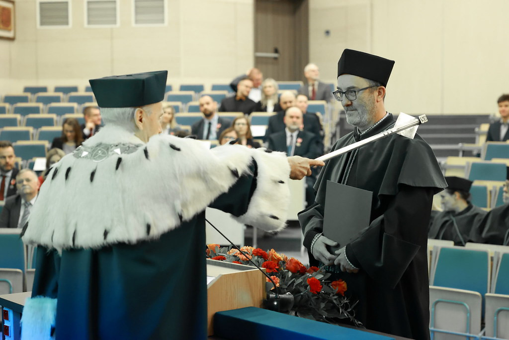 Uroczystość akademicka. Dwóch mężczyzn w togach i biretach. Jeden przekazuje drugiemu berło.
