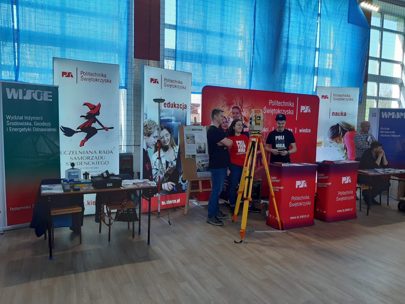 Stoisko Politechniki Świętokrzyskiej z geodetą i banerami. Kluczowe obiekty: geodeta, banery, stół.