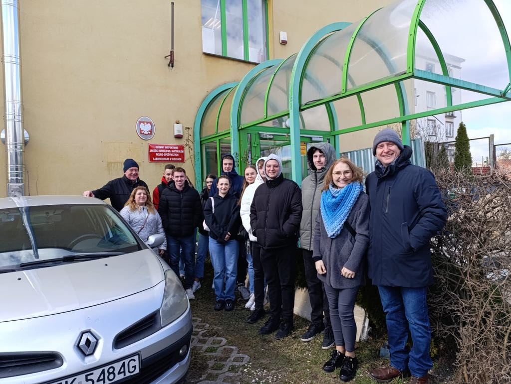 Grupa osób stoi przed budynkiem. Widać samochód, wejście z zieloną konstrukcją i tablicę.