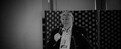 Mężczyzna w okularach przemawia do mikrofonu. W tle widoczna jest zasłona z wzorem. Kontekst: konferencja, spotkanie.